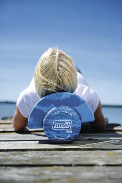 Frau nutzt blauen Sissel Pilates-Roller zur Rückenentspannung am Seeufer.