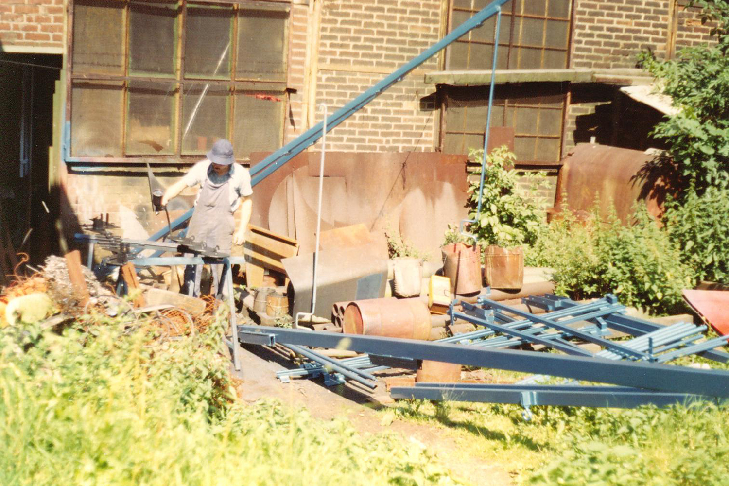 Ein Mann arbeitet in einem alten, an einem Gebäude angebrachten Werkstattraum mit Metallrohren und Werkzeugen, umgeben von Pflanzen und alten Metallteilen.