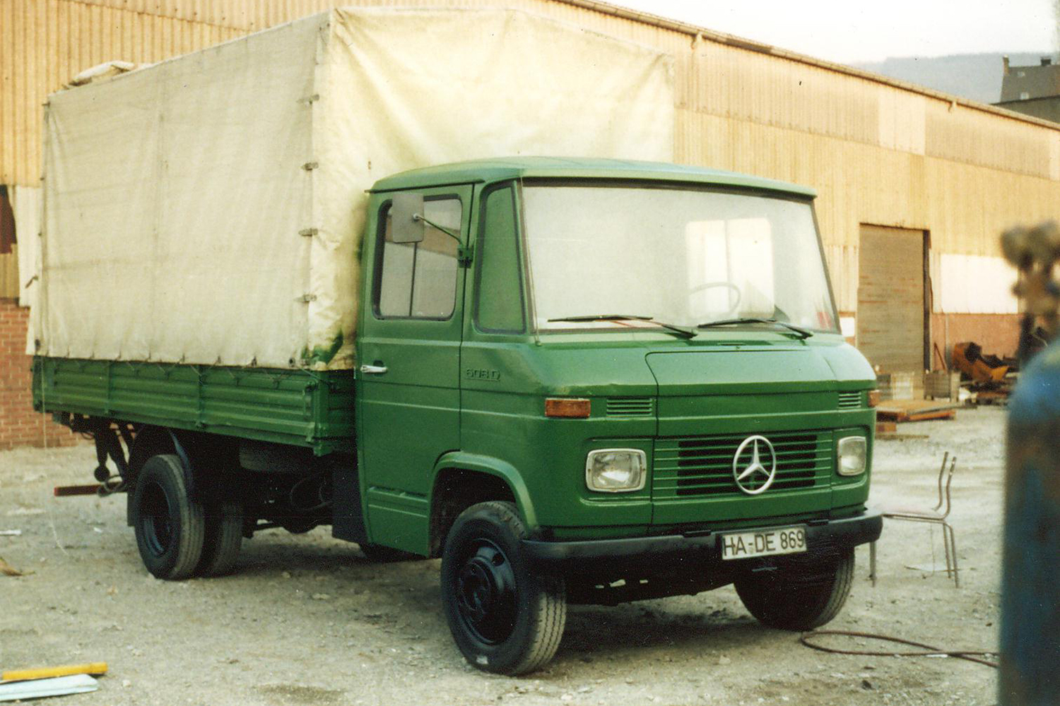 Grüner Mercedes-Benz Lkw mit weißem Planenaufbau, vor einer Lagerhalle.