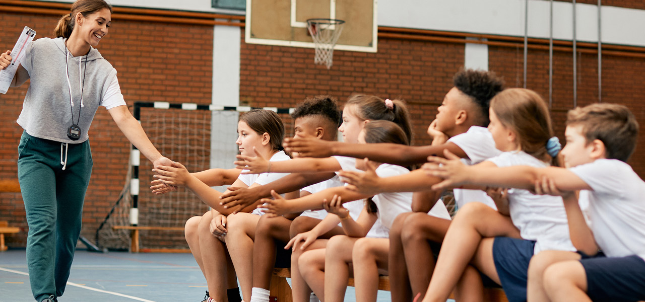 Eine lächelnde Sportlehrerin geht in einer Sporthalle an einer Reihe von Kindern vorbei, die auf einer Bank sitzen, und gibt jedem ein High-Five.