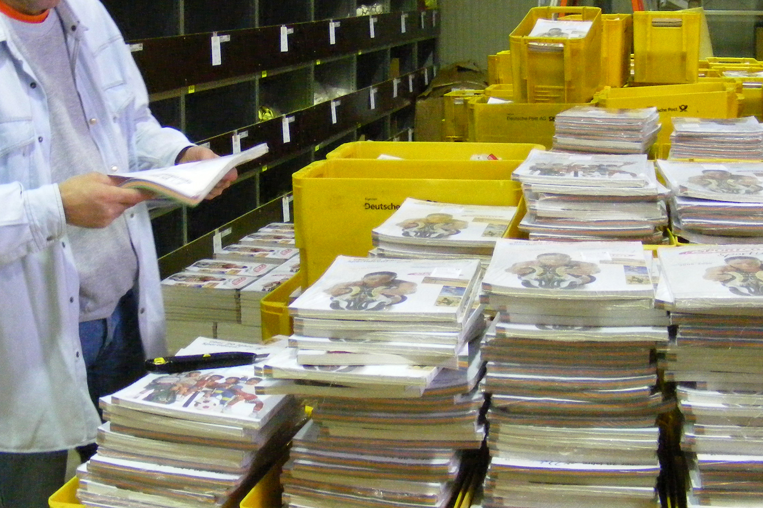 Mehrere Stapel Kataloge in gelben Kisten, eine Person liest einen Katalog in einem Lager.