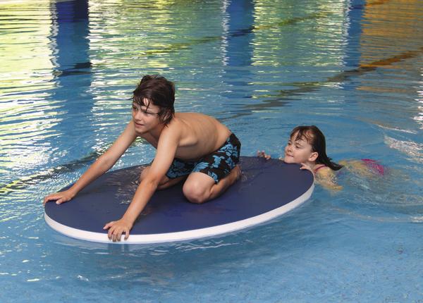 Junge auf Schwimmbrett im Hallenbad, Mädchen schwimmt dahinter, Wasserspaß im Pool.