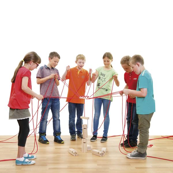 Sechs Kinder spielen kooperativ mit roten Seilen und stapeln Holzklötze auf Holzboden.