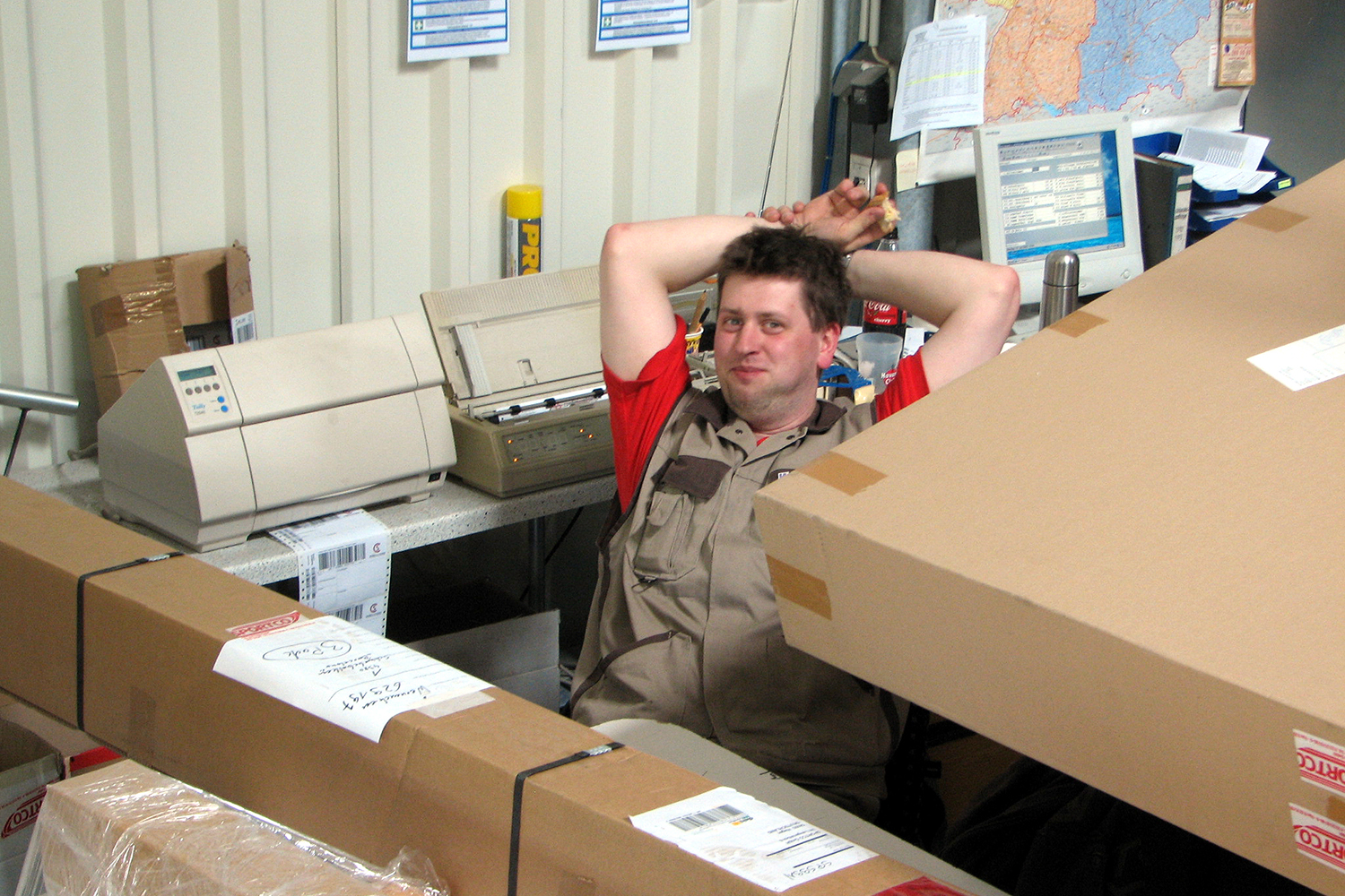 Mann sitzt entspannt mit den Armen hinter dem Kopf auf einem Stuhl in einem Lagerraum, umgeben von Kartons, Verpackungsmaterial und Computern, die für die Bearbeitung von Postsendungen genutzt werden.